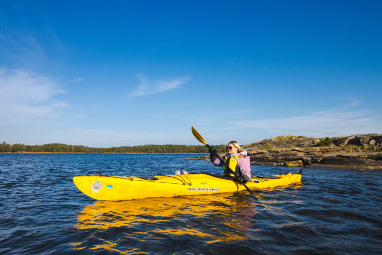 Person in a pink jacket paddling a yellow kayak on a calm body of water, with a rocky shoreline and trees in the background under a blue sky.