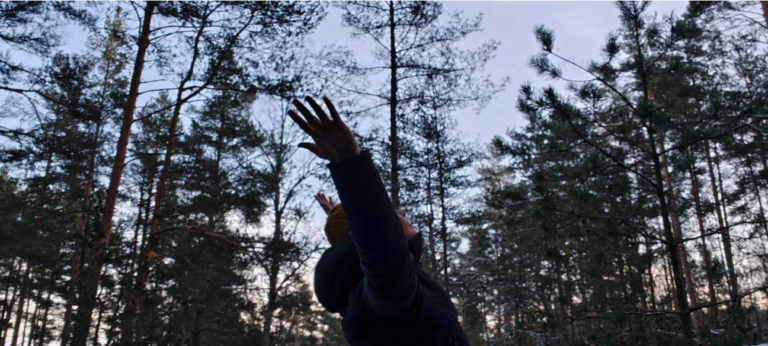 A person in a dark jacket stands in a forest, reaching upward with both arms stretched toward the sky. Trees and a cloudy sky are visible in the background.
