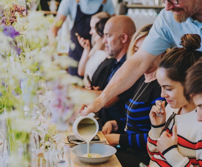 A person pours soup into a bowl on a table lined with people. The table is decorated with flowers and various bowls and utensils.