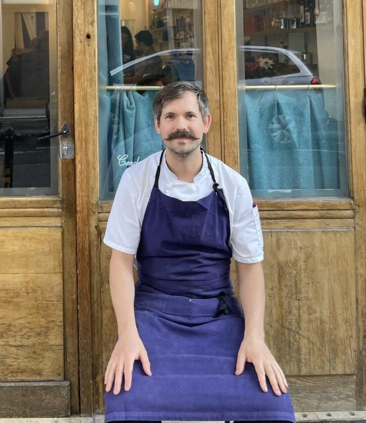 A man in an apron sits on a bench outside a restaurant with wooden doors and blue curtains. The signage above the door reads "RECOIN" and "RESTAURANT".