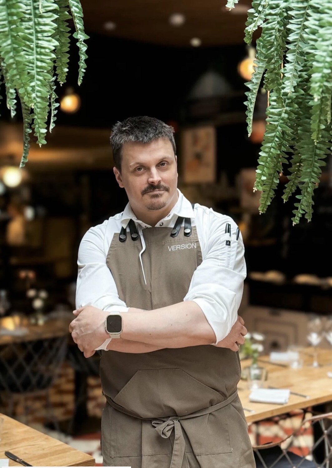 A man in a white shirt and brown apron stands with arms crossed in a cozy, warmly-lit restaurant. Greenery hangs from above.