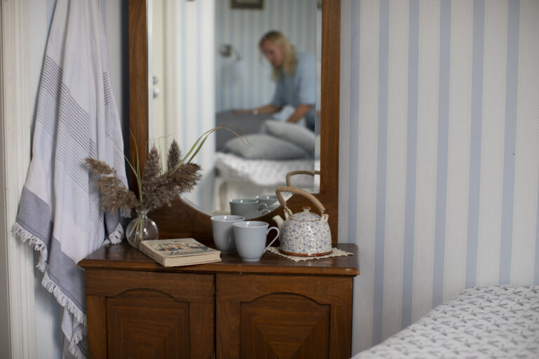 A wooden dresser with three mugs, a teapot, a book, and dried flowers in a vase. A person is blurred in the background, adjusting a bed in a room with striped wallpaper.