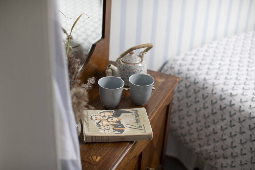 A wooden bedside table with a patterned teapot, two gray mugs, and a book titled "Launched". A patterned bedspread is partially visible in the background.