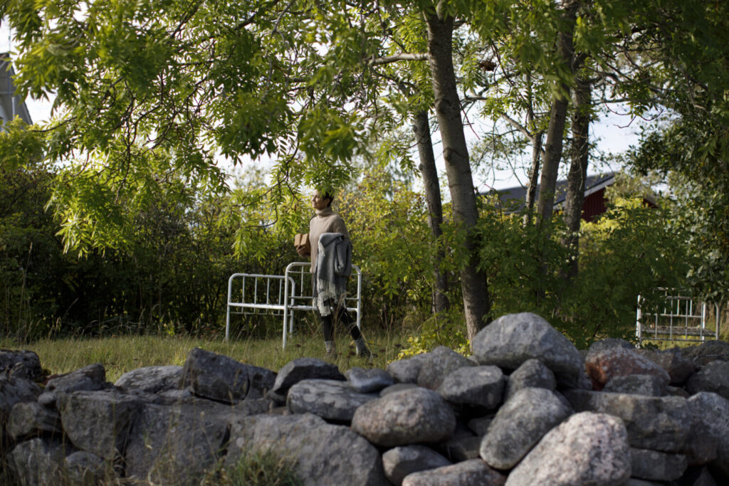 A person carries a metal bed frame under trees, standing near a stone wall in a grassy area.