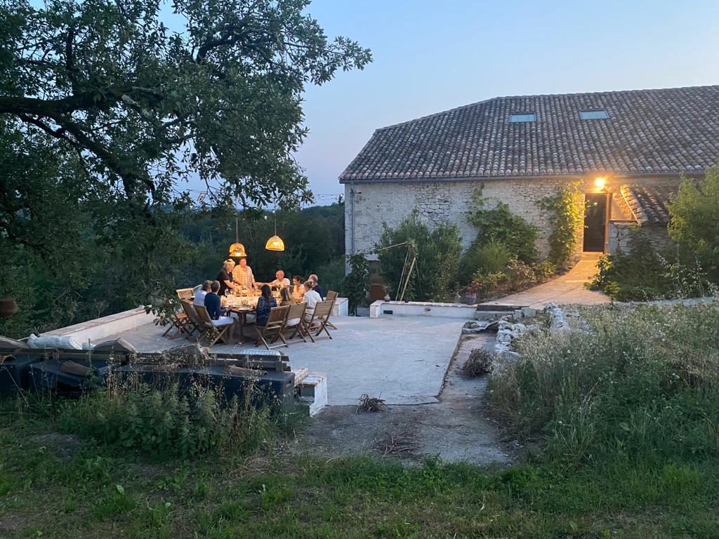 People dining at a table on a rustic outdoor patio near a stone house surrounded by greenery at dusk.