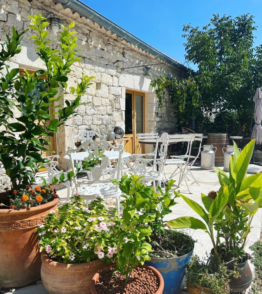 A sunny patio with white metal chairs and tables, surrounded by potted plants and flowers, next to a stone building.