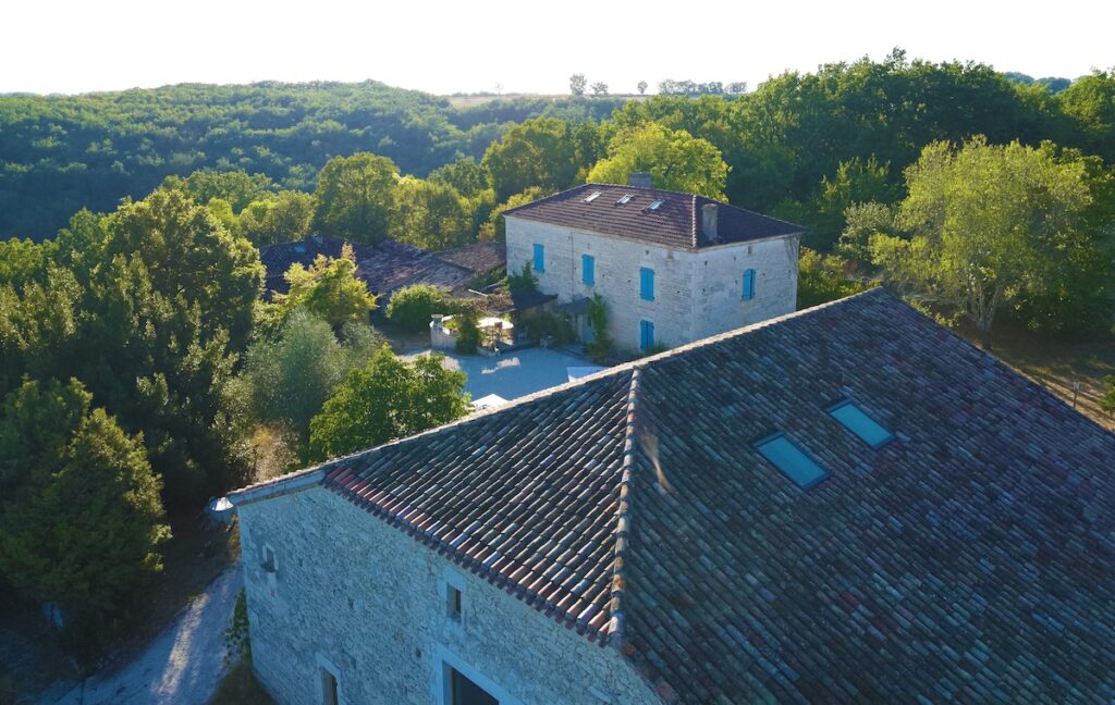 Aerial view of a stone house with a sloped roof surrounded by trees in a rural setting.