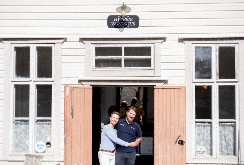 Two people stand smiling and holding hands in front of a building with a sign reading "HYPPEIS VÄRDSHUS." The entrance doors are open, revealing a glimpse of the interior.