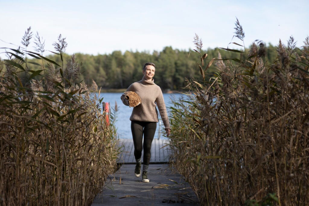 Person in a brown sweater carrying a woven basket, walking on a wooden dock surrounded by tall grasses and a lake in the background.