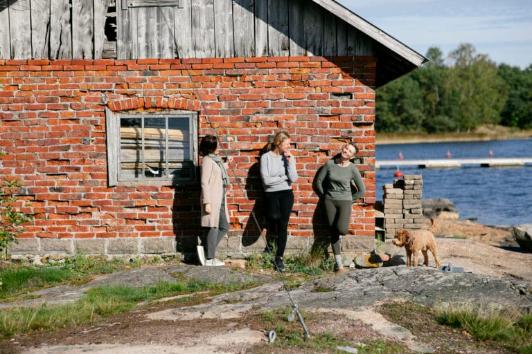 Three people standing by a brick building near a body of water, with a small dog nearby on a rocky surface.