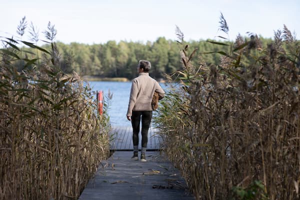 A person in a cozy sweater strolls along a wooden path toward a serene lake, surrounded by Finland's tall grass. A forest provides the perfect backdrop, reminiscent of an archipelago retreat, promising tranquility and perhaps the warmth of a nearby sauna.