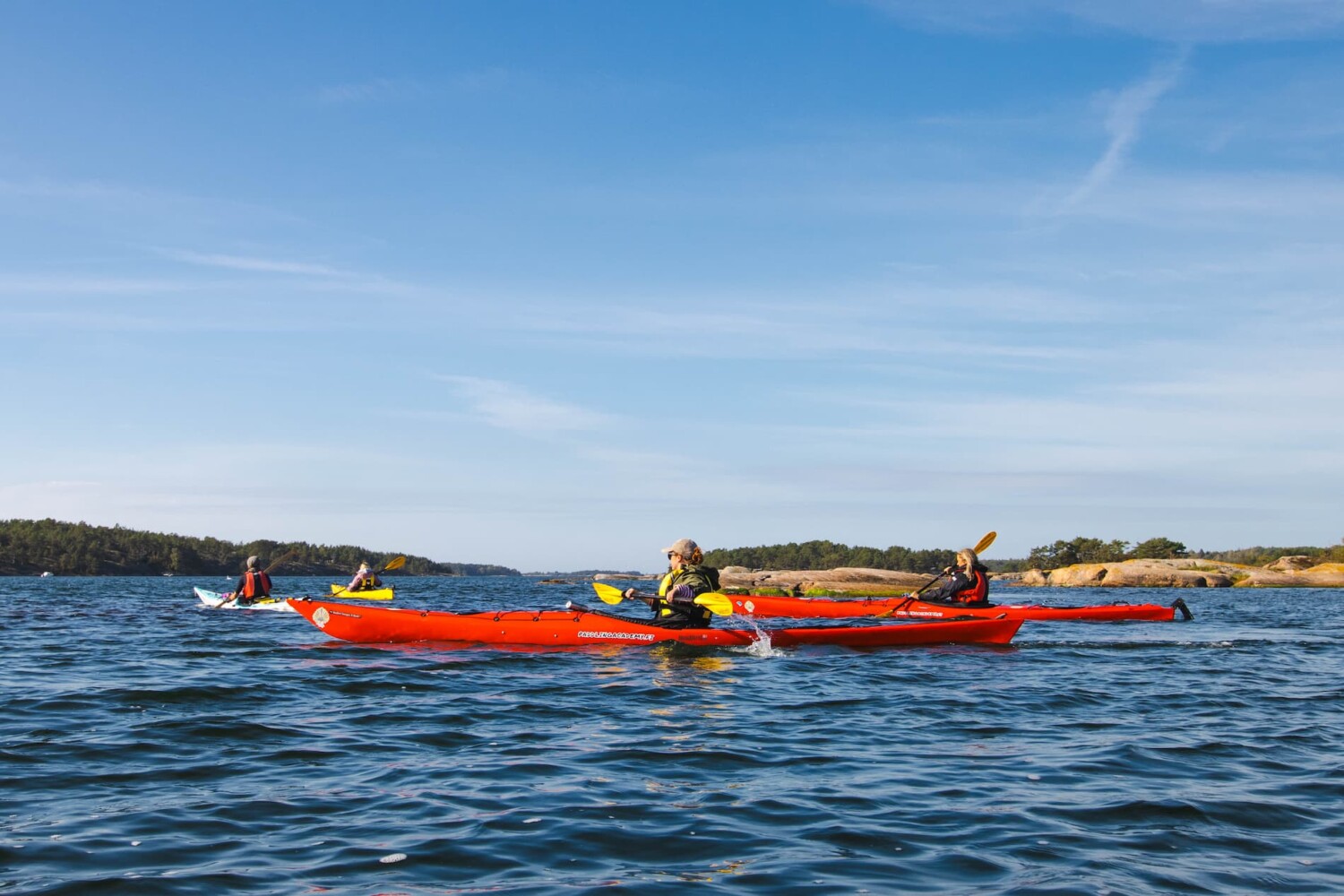 Three people kayaking on a calm body of water under a clear blue sky, with rocky islands visible in the background.