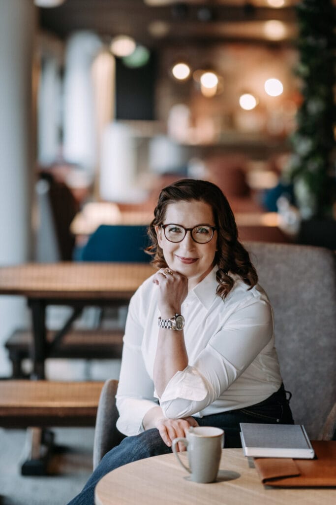 A person wearing glasses and a white shirt sits at a table with a coffee cup and notebook, resting their chin on their hand in a cozy cafe setting.