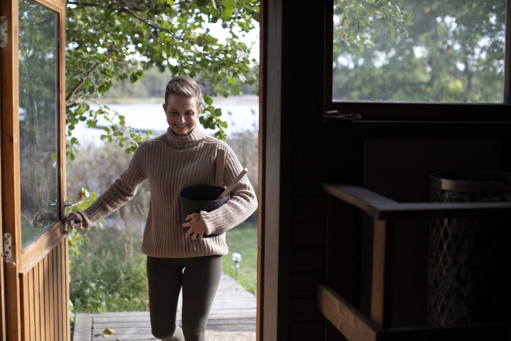 A person wearing a beige sweater enters a wooden cabin, holding a black pot. Trees are visible in the background.