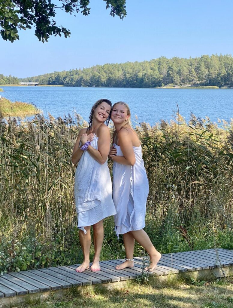 Two people stand on a wooden path near a tranquil body of water in Finland's archipelago, wrapped in towels after a refreshing sauna, with tall grass and trees framing the scene under a clear blue sky.