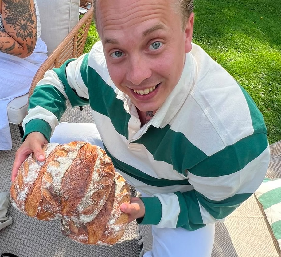 A person in a green and white striped shirt smiles while holding two loaves of rustic bread outdoors on a sunny day.